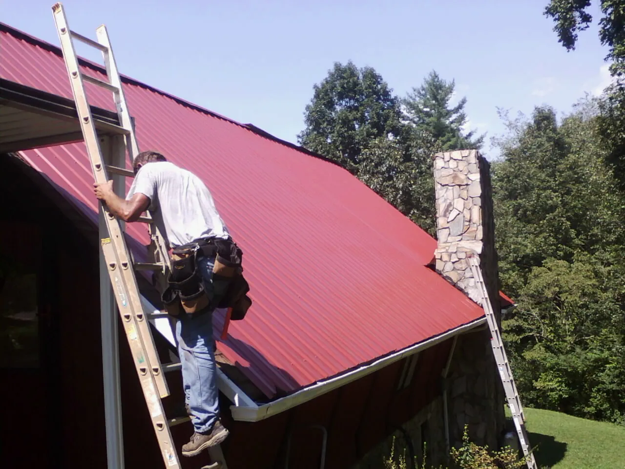 Roofer on a ladder beside a red metal roof and stone chimney — Soffit & Fascia Repair by Roofing Brothers in Hanson